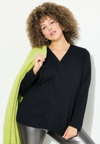 Schwarzes Langarm-Oberteil mit tiefem V-Ausschnitt; weicher Stoff, lockere Passform. Das Model trägt glänzende silberne Leggings und wirft sich einen strukturierten, leuchtend grünen Schal um.