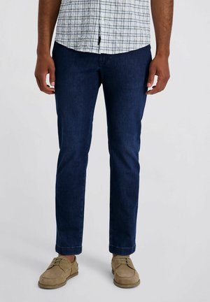 Man draagt donkere blauw slim-fit jeans, beige casual schoenen en een wit kortademig shirt met een blauw ruitpatroon.