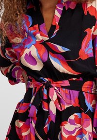 Robe portefeuille fleurie en tissu noir ornée de grands motifs floraux rouges, roses et bleus. Taille nouée avec un nœud, manches 3/4 avec poignets froncés.