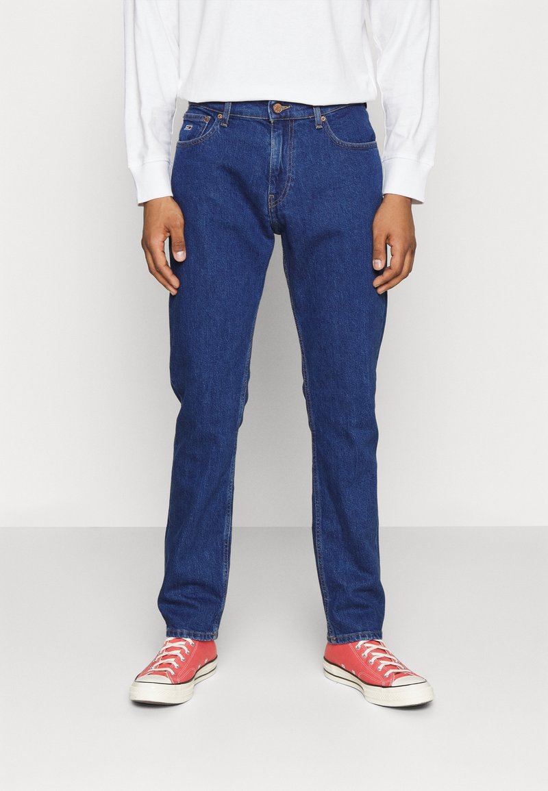 Jeans in denim blu con vestibilità dritta, cinque tasche e zip. Indossati con scarpe da ginnastica rosse, con lacci bianchi e suola in gomma.