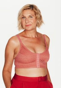 Bralette en dentelle rose avec une fermeture à l'avant, des bretelles larges et des motifs texturés. Porté avec un pantalon rouge taille haute.