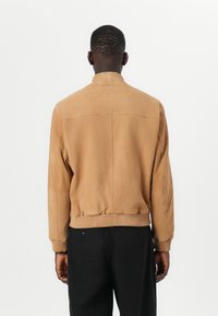 Béžová semišová bomber jacket s rebrovaným golierom a manžetami. Má priliehavý pás, hladkú textúru a minimálne prešívanie.