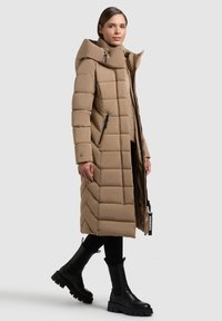 Dlouhý béžový prošívaný kabát s vysokým límcem, zdobený vertikálním a rybinovým vzorem prošívání, se dvěma bočními kapsami na zip a hladkým povrchem.