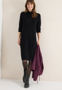 Schwarzes geripptes Strickkleid mit langen Ärmeln, hohem Kragen und Kordelzug. Schwarze Schnürstiefel und eine kastanienbraune Jacke.