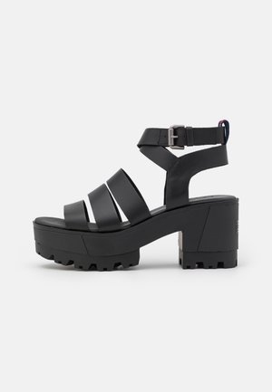 Sort sandaler med chunky hæl, tre brede remme foran, ankelrem med spænde og robust gummisål på hvid baggrund.
