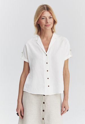 Mujer con cabello rubio hasta los hombros, que lleva una camisa blanca de manga corta con botones y una falda beige con botones delanteros.