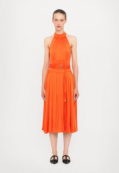 Vestido naranja sin mangas con falda plisada y cintura ceñida que presenta una hebilla circular. Textura suave, hasta la rodilla. Zapatos planos negros.