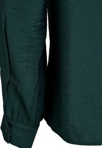 Polsino di camicia a maniche lunghe verde scuro con chiusura a bottone e tessuto leggermente strutturato su sfondo uniforme.