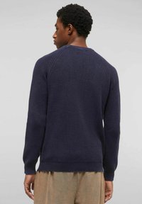 Uomo che indossa un maglione a maglia blu scuro e pantaloni beige, rivolto di spalle contro uno sfondo grigio chiaro uniforme.