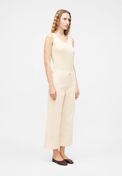 Γυναίκα με μακριά κυματιστά μαλλιά, ντυμένη με αμάνικη κρεμ μπλούζα, φαρδιά μπεζ παντελόνια με μανσέτες και σκούρα καφέ επίπεδα παπούτσια, στέκεται μπροστά σε λευκό φόντο.