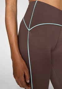 Brune atletiske tights med lyseblå kontrastsøm, høy midje design og et teksturert stoff. Detaljerte sømmer gir økt struktur.