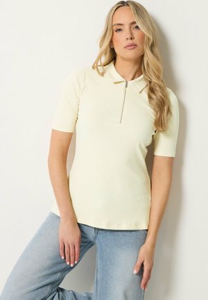 Donna bionda che indossa una polo crema a maniche corte a coste con zip frontale, abbinata a jeans azzurri chiari, in piedi contro una parete bianca.