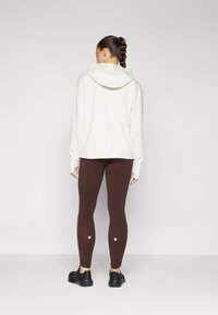 Sudadera blanca con capucha y mangas largas, combinada con leggings marrones que tienen acentos circulares en la parte inferior de las piernas, y zapatillas deportivas negras.