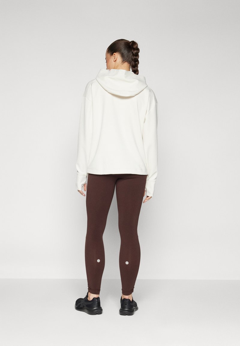 Witte hoodie met een capuchon en lange mouwen, gecombineerd met bruine legging met cirkelaccenten op de onderbenen, en zwarte sportschoenen.