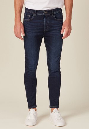 Donkerblauwe slim-fit jeans met een gladde denimtextuur, vijf zakken en subtiele vervaging. Gecombineerd met witte sneakers voor contrast.