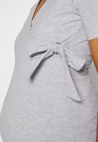 Tät bild av en grå rippad graviditetstop med en sidoknut, som bärs av en gravid person.