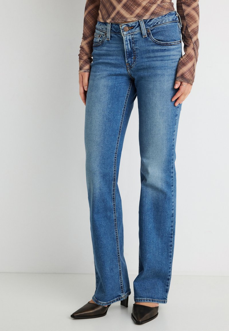 Γυναίκα που φοράει μπλε τζιν με ίσια παντελόνια, μαύρα μυτερά τακούνια και καφέ μακρυμάνικο πουκάμισο με καρό σχέδιο, στέκεται μπροστά σε απλό λευκό φόντο.