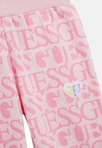Pantalons de survêtement rose clair avec un motif "GUESS" intégral. Doté d'une ceinture rose texturée et d'un patch logo en forme de cœur.
