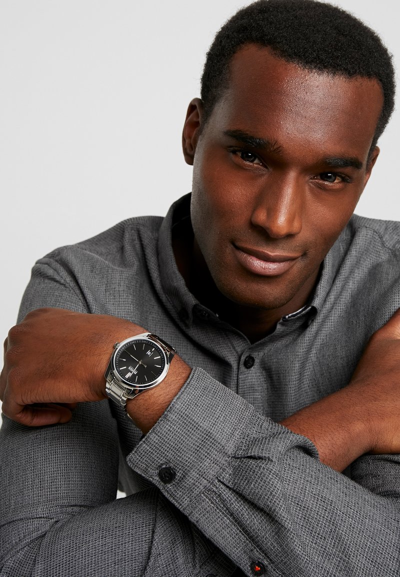 Men's watch with a black face, silver-tone stainless steel bracelet, and a textured grey shirt. Features include a round case and clear markings.