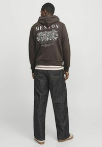 Brauner Kapuzenpullover mit "MENTON" Grafik und Gebäuden auf dem Rücken, kombiniert mit locker sitzenden dunklen Jeans und beigen Sneakern. Grobe Textur sichtbar.