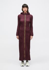 Bordeauxroter, gesteppter Mantel mit Kapuze, ausgestattet mit einem durchgehenden gelben Reißverschluss und strukturierter Oberfläche. Längere Silhouette, kombiniert mit hellfarbenem Schuhwerk.