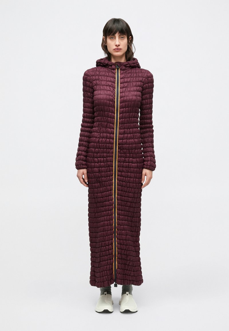 Bordeauxroter, gesteppter Mantel mit Kapuze, ausgestattet mit einem durchgehenden gelben Reißverschluss und strukturierter Oberfläche. Längere Silhouette, kombiniert mit hellfarbenem Schuhwerk.