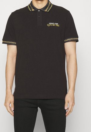 Schwarzes Poloshirt mit gelbem Kettenbesatz am Kragen und an den Ärmeln, mit dem "Versace Jeans Couture" Logo auf der Brust, getragen von einer stehenden Person.