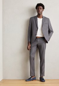 Costum gri cu o croială slim, jachetă cu un rând de nasturi, tricou alb și mocasini din suede albastru. Textură netedă, design minimalist, curea maro.