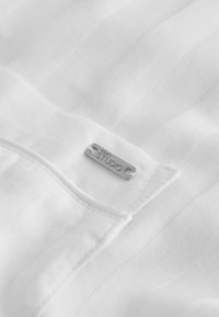 Tissu blanc avec des fines rayures verticales, doté d'une petite étiquette métallique rectangulaire gravée « NEXT STUDIO » près d'une couture.