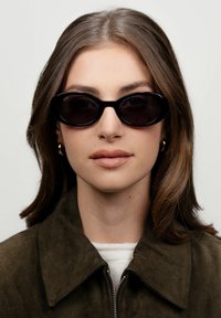 Černé oválné sluneční brýle s tmavými skly, silnými obrubami a hladkým povrchem. Nosí je modelka s hnědými vlasy, v tmavě zelené semišové bundě.