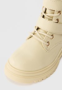 Bota de couro creme com bico arredondado, várias posições para os atacadores e ilhós em tom dourado; sola texturizada com um padrão de ranhura robusto.