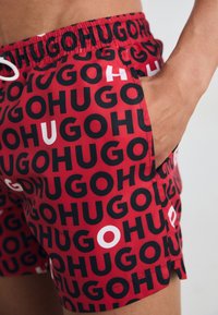 Rote Badehose mit einem sich wiederholenden schwarzen und weißen "HUGO"-Muster, elastischem Bund und seitlichen Taschen.