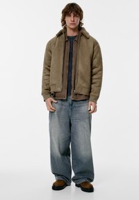 Hnědá bomber bunda s kožíškovým límcem, vrstvená přes texturovaný šedý svetr, doplněná světle modrými širokými džíny a hnědými botami.