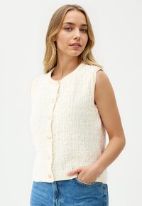 Romige textuur gebreide vest, mouwloos ontwerp, ronde halslijn, sluiting met knopen aan de voorkant met gouden knopen, en geribbelde zoom. Geselecteerd met denim shorts.