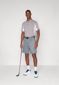 Camisa polo cinza com detalhes brancos, calções cinzas e sapatilhas de golfe brancas. Segurando um taco de golfe, de pé sobre um fundo neutro.