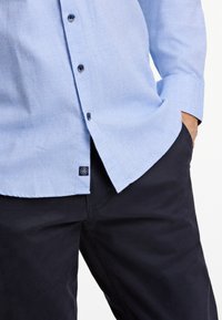 Ljusblå skjorta med kort ärm, mörka knappar och en etikett i nederkanten, som kombineras med mörkblå byxor. Texturerat tyg med en avslappnad passform.