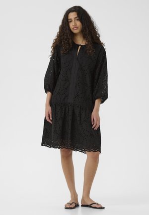 Schwarzes Kleid mit langen Puffärmel, Lochmuster, Rundhalsausschnitt, Schlüssellochdetail und gestuftem Saum. Kombiniert mit schwarzen Flip-Flops.