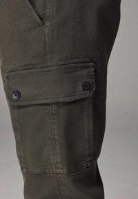 Cargo-Hosen aus olivgrünem Canvas, mit einer seitlichen Tasche mit Klappe und schwarzen Knöpfen, strukturiertem Stoff und sichtbaren Nahtdetails.
