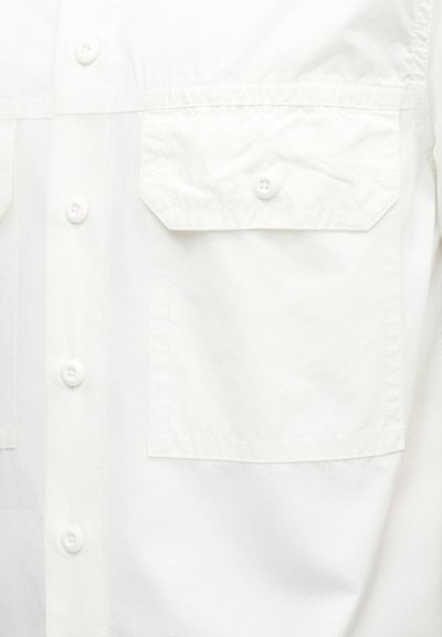 Λεπτομέρεια λευκού πουκάμισου με τσέπη με καπάκι στη δεξιά πλευρά με κούμπωμα με κουμπί και ορατά κουμπιά στο μπροστινό μέρος.