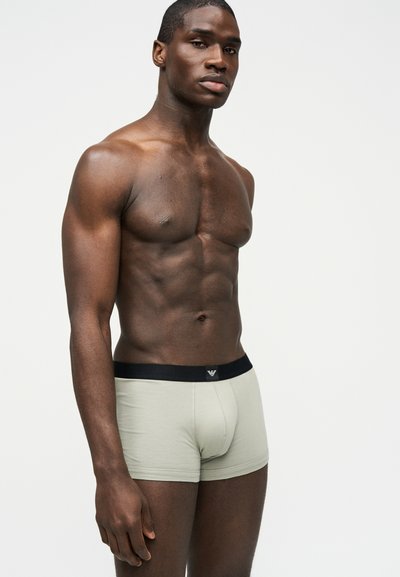 Μυώδης άντρας, όρθιος, φορώντας ανοιχτό γκρι μποξέρ με μαύρη ζώνη, πάνω σε απλό ανοιχτόχρωμο φόντο.