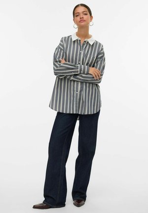 Donna in piedi con le braccia incrociate, indossa una camicia oversize a righe verticali con colletto bianco, jeans larghi scuri, orecchini a cerchio e scarpe a punta.