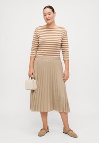 Beige Faltenrock mit einem taillierten Bund, kombiniert mit einem braun-weißen gestreiften Langarmtop. Hält eine kleine weiße Handtasche.