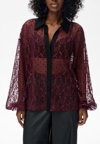 Bordowa przezroczysta bluzka z czarnym kołnierzem, ozdobnym wzorem koronki, półprzezroczystą tkaniną, zapinana na guziki z przodu oraz z bufiastymi rękawami.