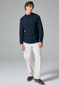 Marineblaue Strickpullover mit strukturierten Zopfmustern, Rundhals-Ausschnitt und gerippten Bündchen. Kombiniert mit cremefarbenen Hosen und dunkelbraunen Schuhen.