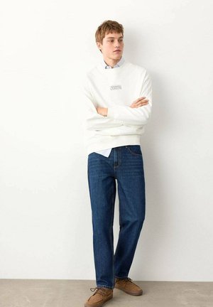 Hanorac alb cu guler rotund și logo frontal, cămașă albastră cu guler dedesubt, blugi drepți de culoare albastru închis și pantofi maro cu șireturi.