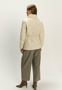 Frau in einer beigefarbenen gesteppte Jacke, karierter weit geschnittener Hose und braunen Wildleder-Stiefeln mit Absatz, die mit dem Rücken zu einer schlichten Hintergrund steht.