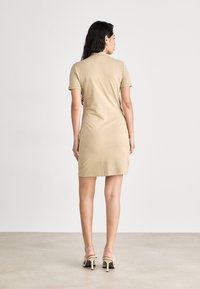 Robe beige légèrement au genou avec un col polo et des manches courtes. Le tissu semble lisse et extensible. Tenue complétée par des talons ouverts.