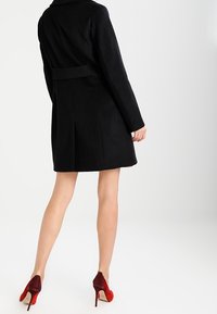 Zwarte wolmixjas met een tailordesign, voorzien van een riemdetail in de taille en een licht uitlopende zoom. Draagt een paar rode hoge hakken.