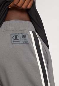 Pelēki svārki ar sānu logotipa iešuvumu, kurā redzami "92" un "AUTHENTIC ATHLETICWEAR". Krāsoti ar melnbaltiem svītrainiem akcentiem sānos.