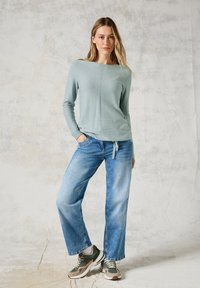 Haut à manches longues en teal clair texturé, associé à un jean large bleu délavé et des baskets vertes, se tenant contre un fond gris clair.